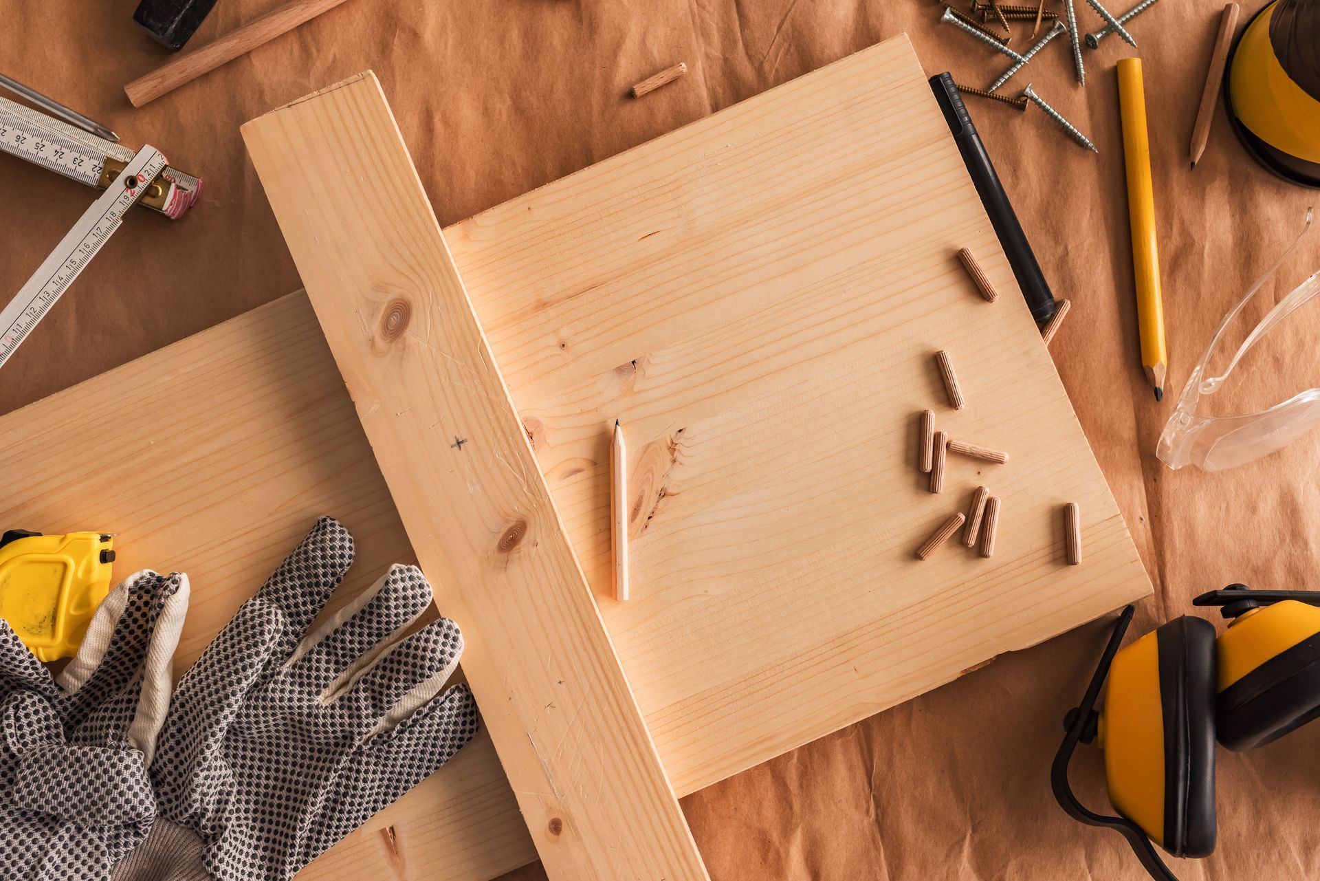 Top-down view of wood planks on a brown surface with a carpenter's square, wooden dowels, gloves, and safety gear.