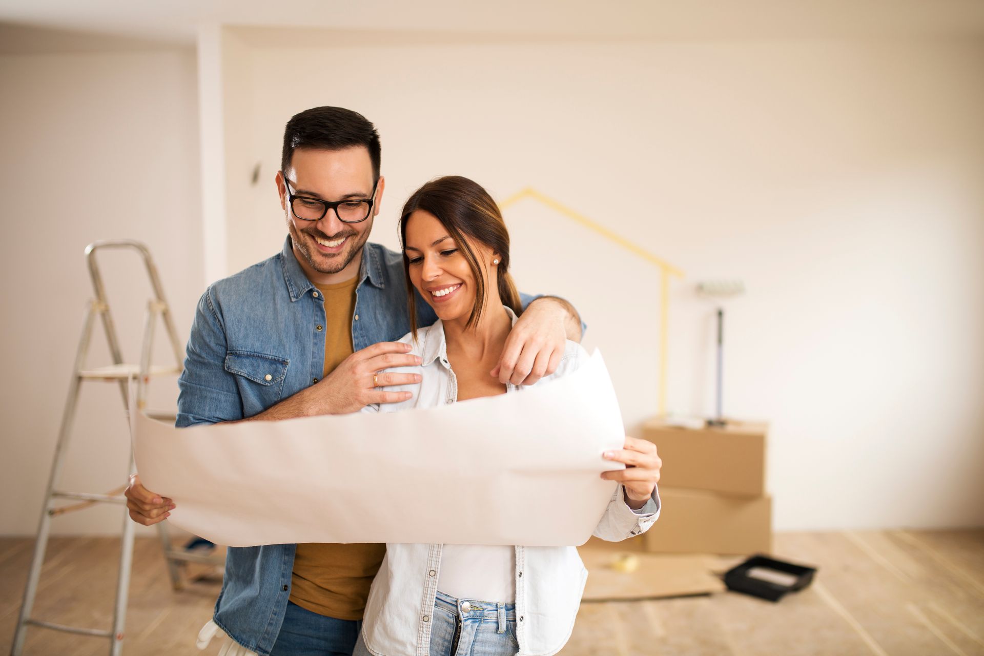 A couple smiles while looking at architectural plans inside an unfurnished room with a ladder and cardboard boxes.