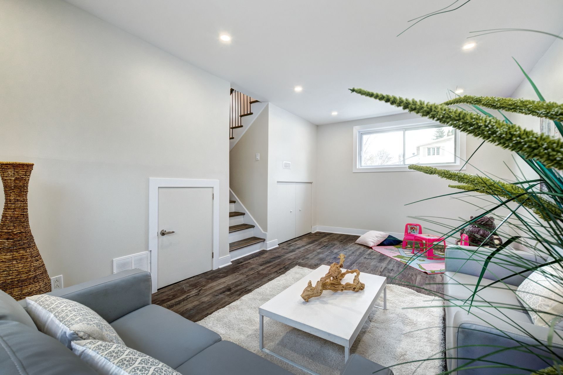 A modern living room with a gray sofa, white coffee table, and area rug, featuring stairs and a window in the background.