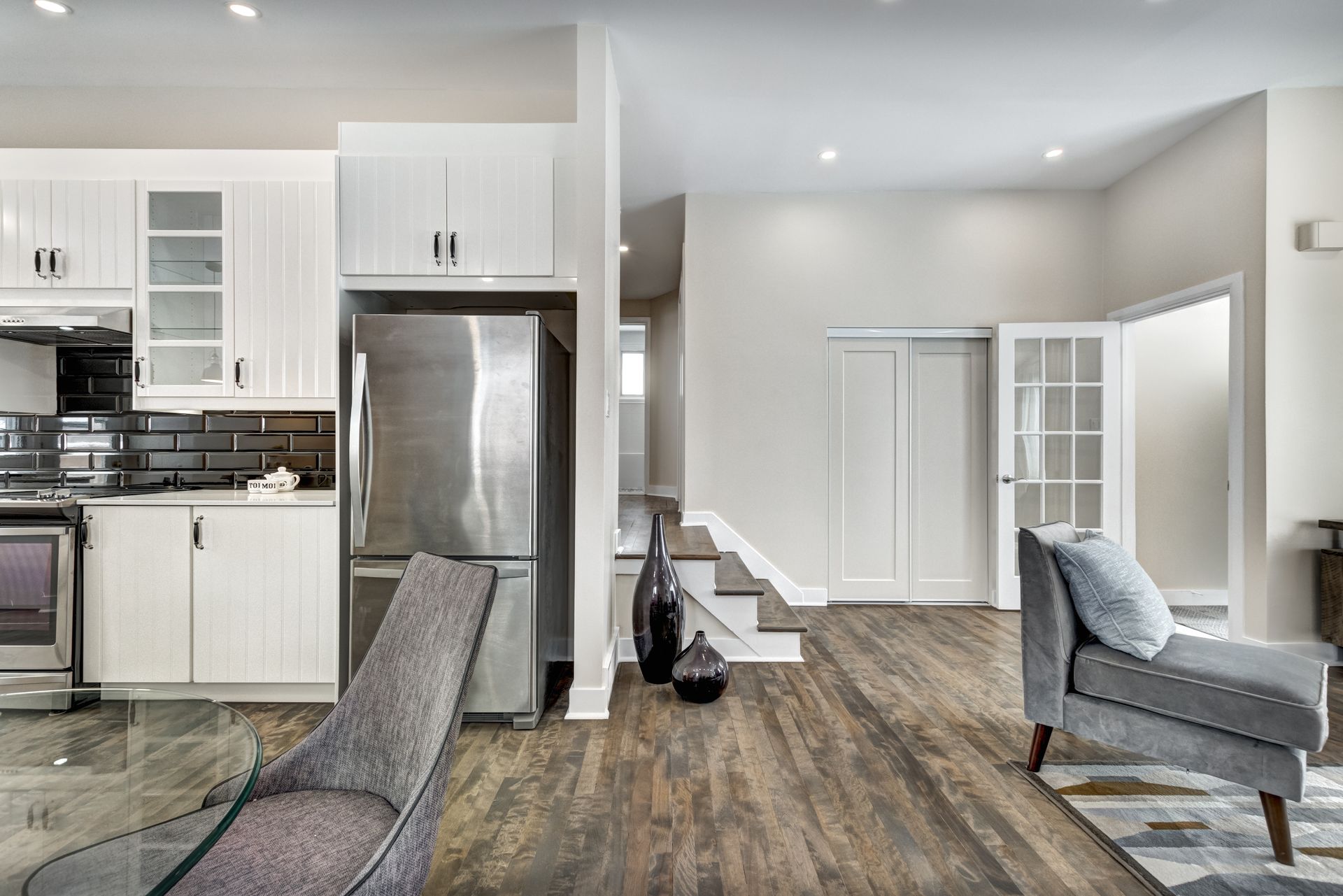 An open kitchen and living area featuring white cabinets, stainless steel appliances, wood floors, and gray armchairs.