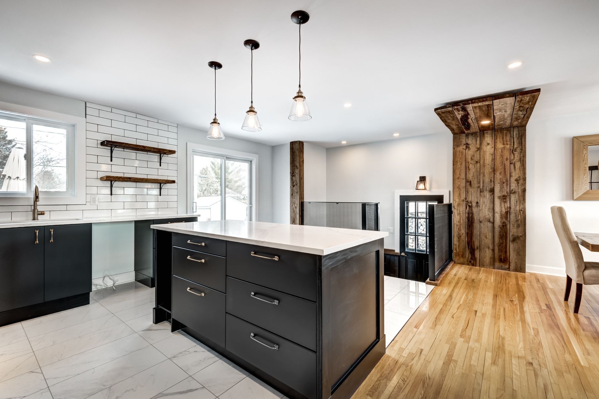 Modern kitchen with dark cabinets, a white island, white tiled floor, and a rustic wooden accent wall with a staircase.