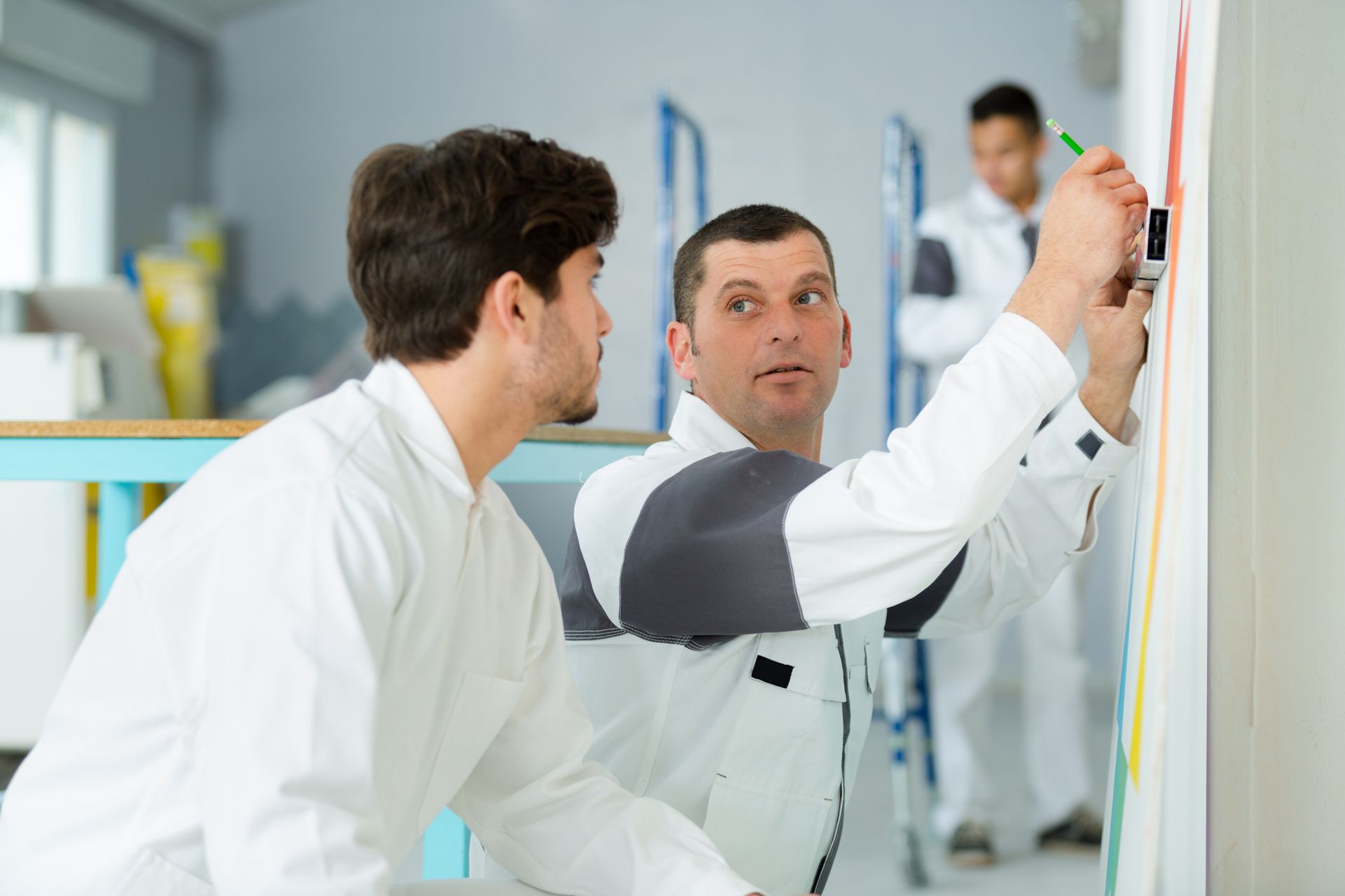 Two technicians in white uniforms working on a project, with one marking a measurement on a wall and another observing.