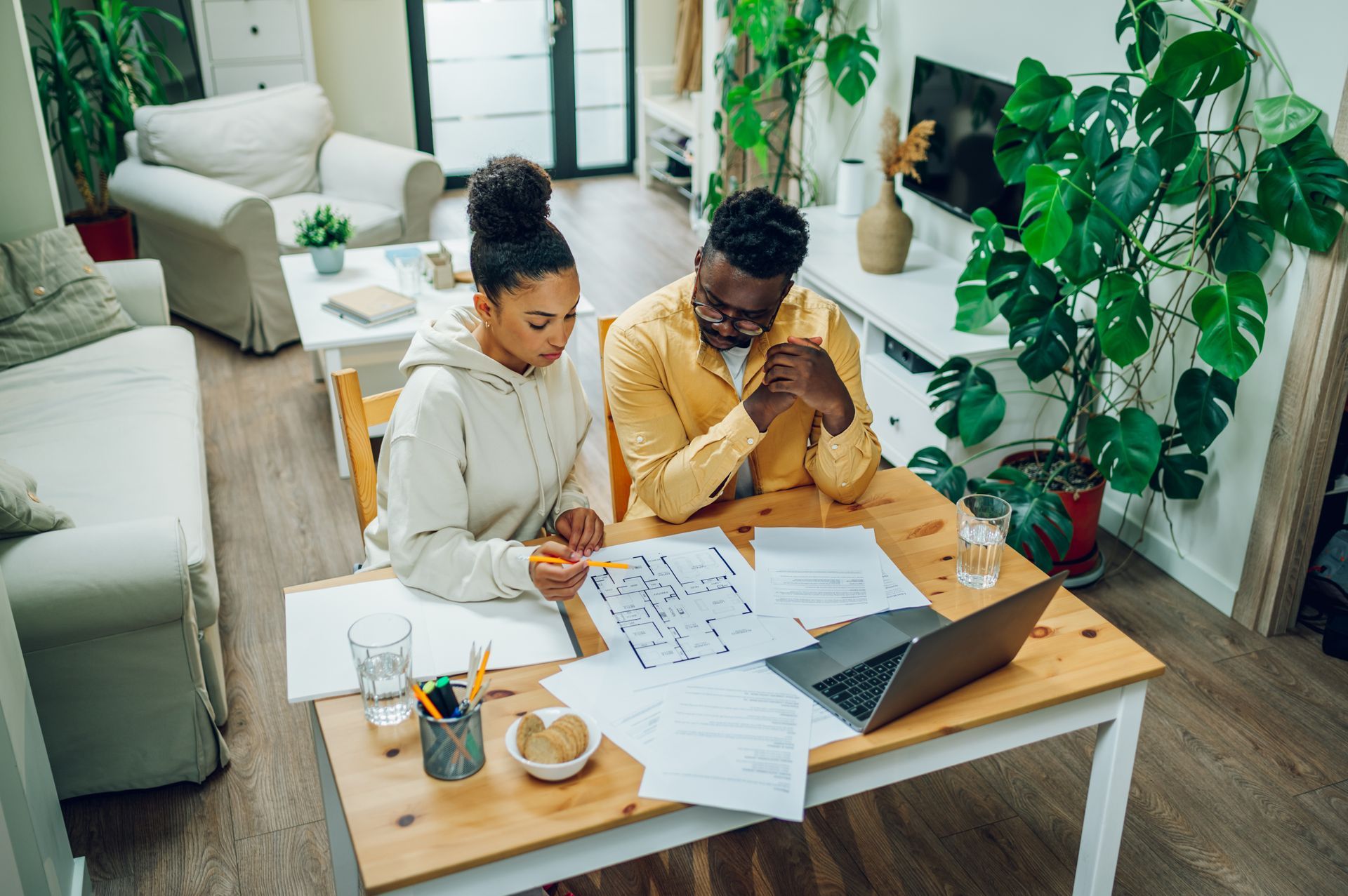 Two people sit at a wooden table in a bright, plant-filled room, reviewing architectural floor plans and a laptop.