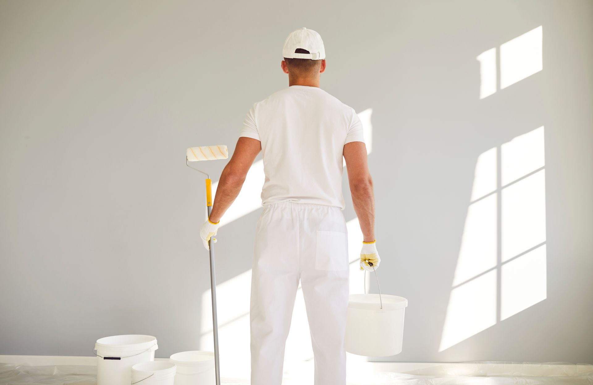 A person in white work clothes stands with a paint roller and bucket, facing a freshly painted light gray wall.