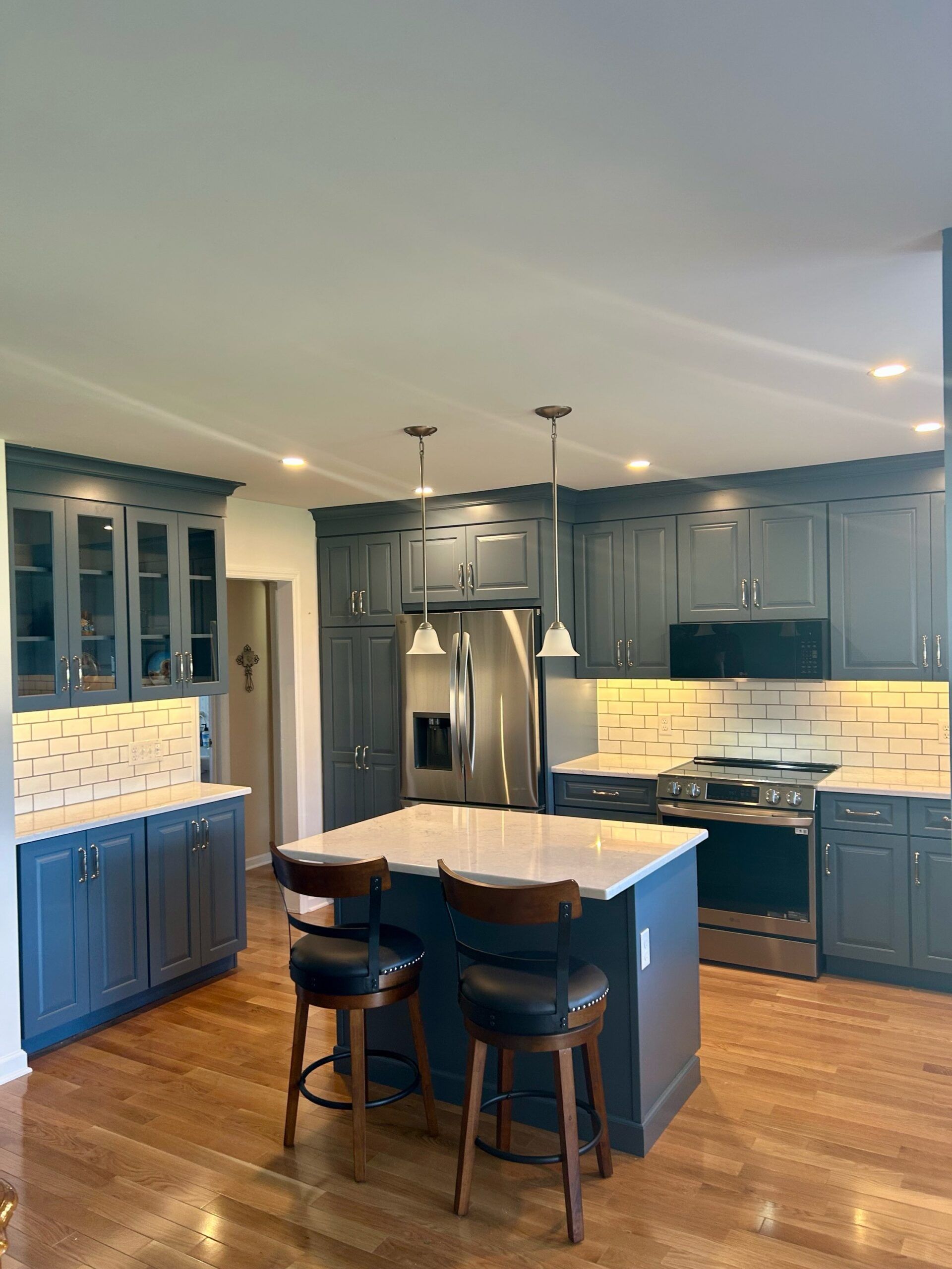 A kitchen with blue cabinets, a white-topped island with two stools, stainless steel appliances, and hardwood floors.