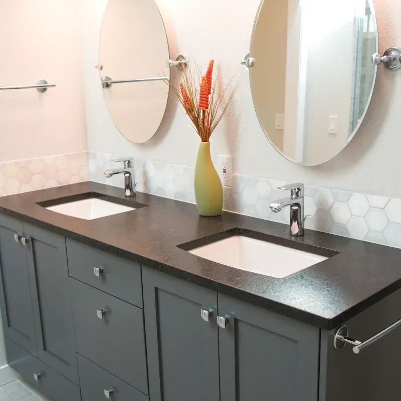 Double vanity with a dark countertop, grey cabinets, two oval mirrors, and white hexagonal tile backsplash.