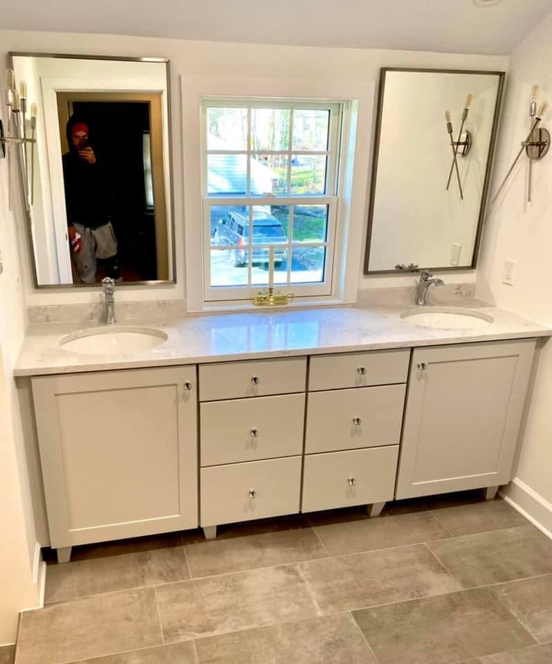 A bathroom vanity with dual sinks, a center drawer unit, and two mirrors flanking a window, featuring grey cabinets.