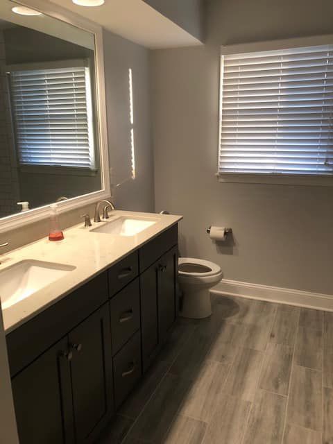 A modern bathroom featuring a dark vanity with a white countertop, large mirror, and a toilet under a window with blinds.