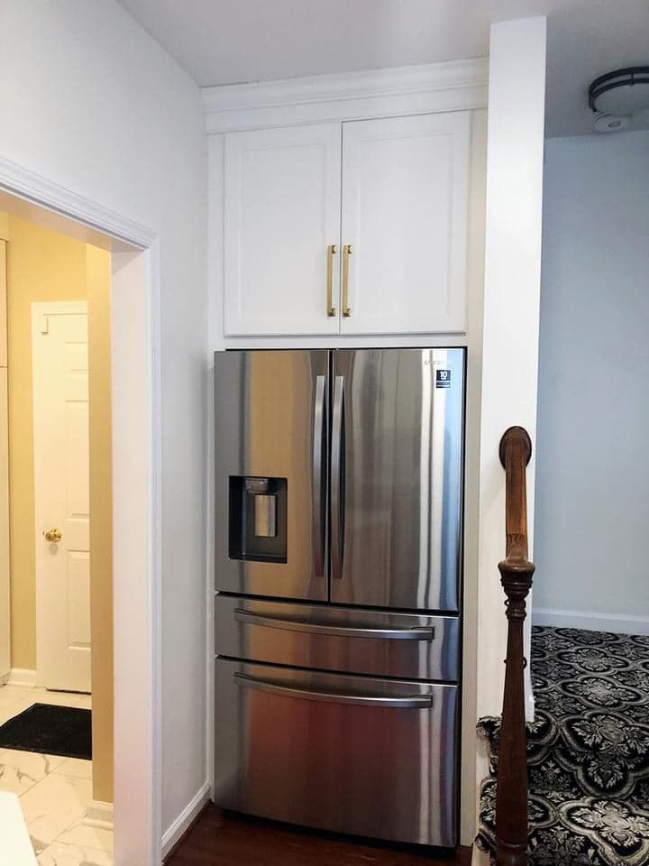 A stainless steel refrigerator sits in a recessed alcove beneath white cabinets with brass handles next to a staircase.