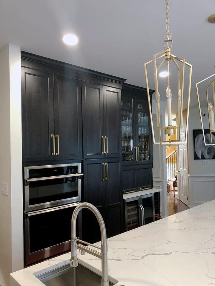 A modern kitchen featuring black cabinetry, gold hardware, a double oven, a wine cooler, and a white marble island.