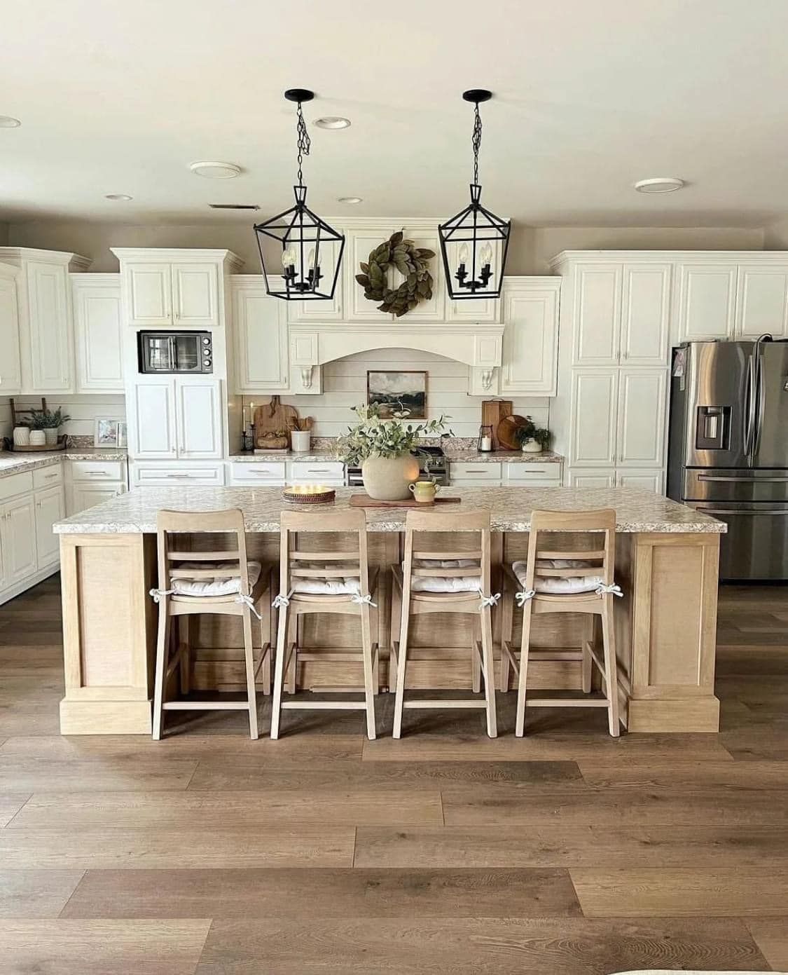 A bright, farmhouse-style kitchen with a wooden island, white cabinetry, two hanging lanterns, and stools.