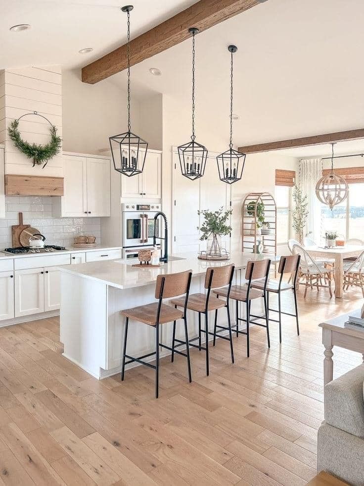 A bright modern kitchen with a white island, three pendant lights, wood beams, and a dining area in the background.