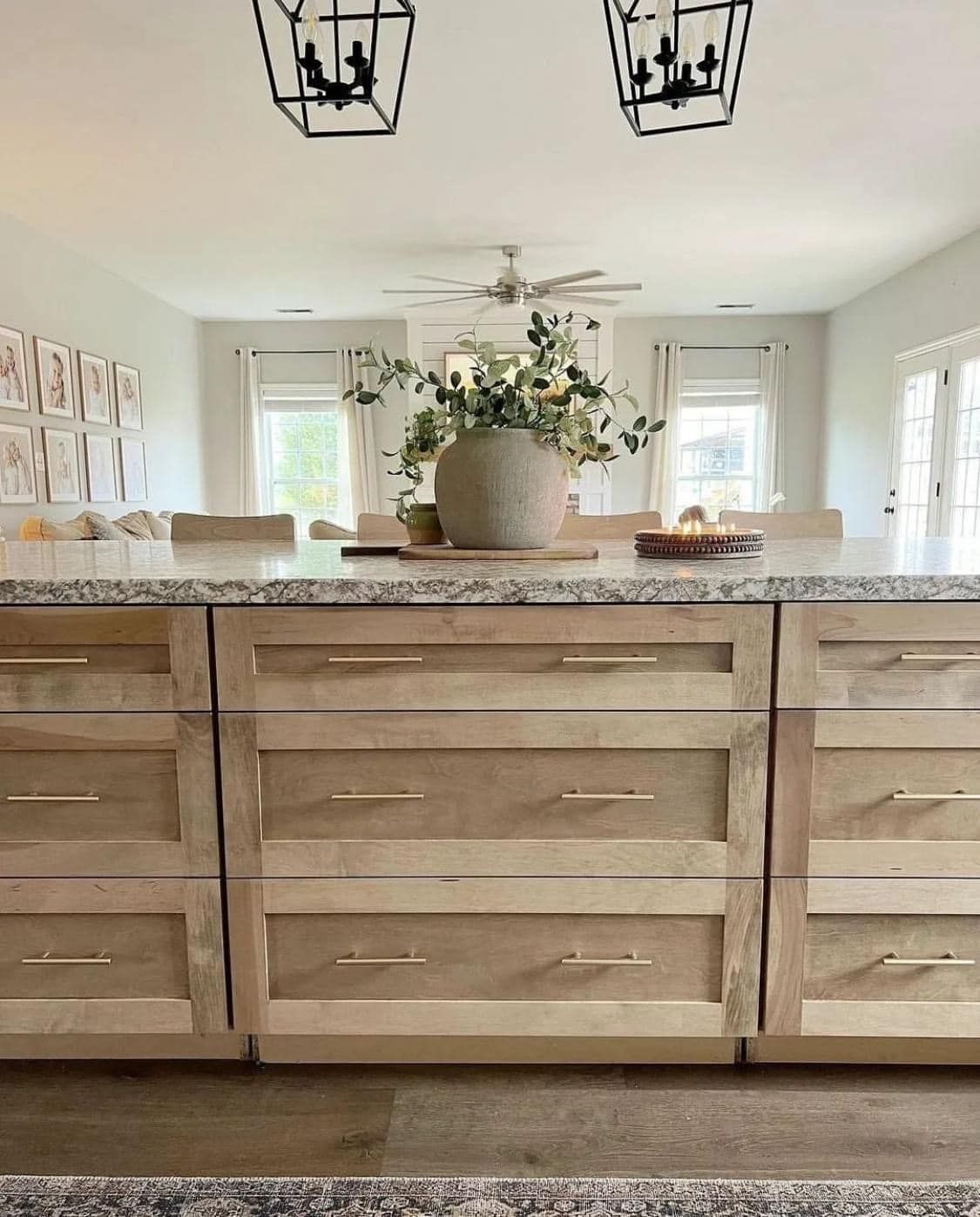 A wood-toned kitchen island with multiple drawers sits in a bright, modern room with hanging lanterns and greenery.