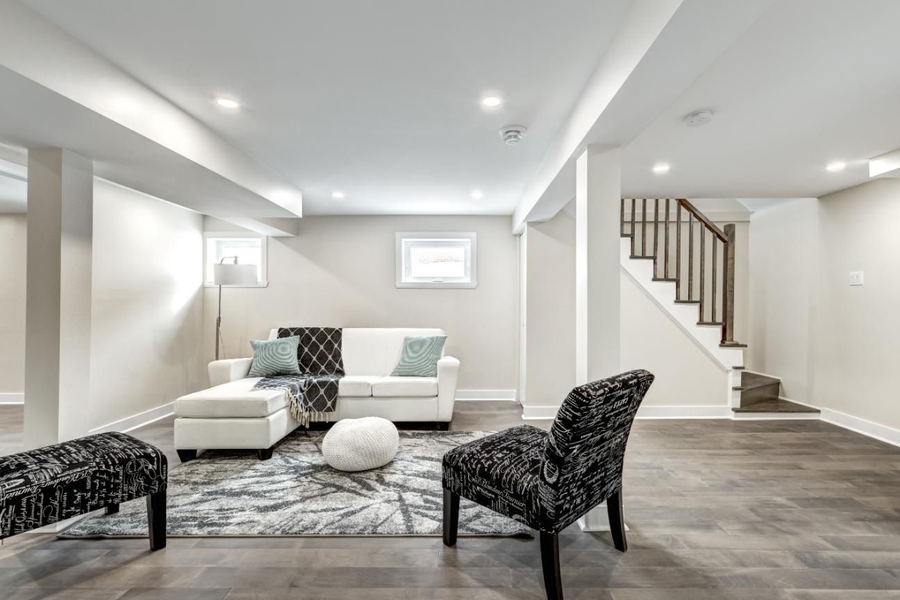 A bright basement living room with a white sectional, patterned chairs, a patterned rug, and a staircase.