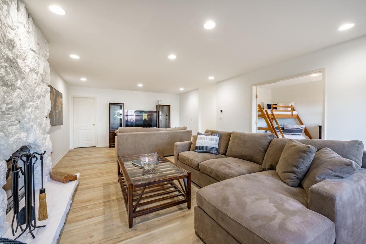 A living room with a stone fireplace, beige sectional sofa, glass-topped coffee table, and a view into a bunk bed room.
