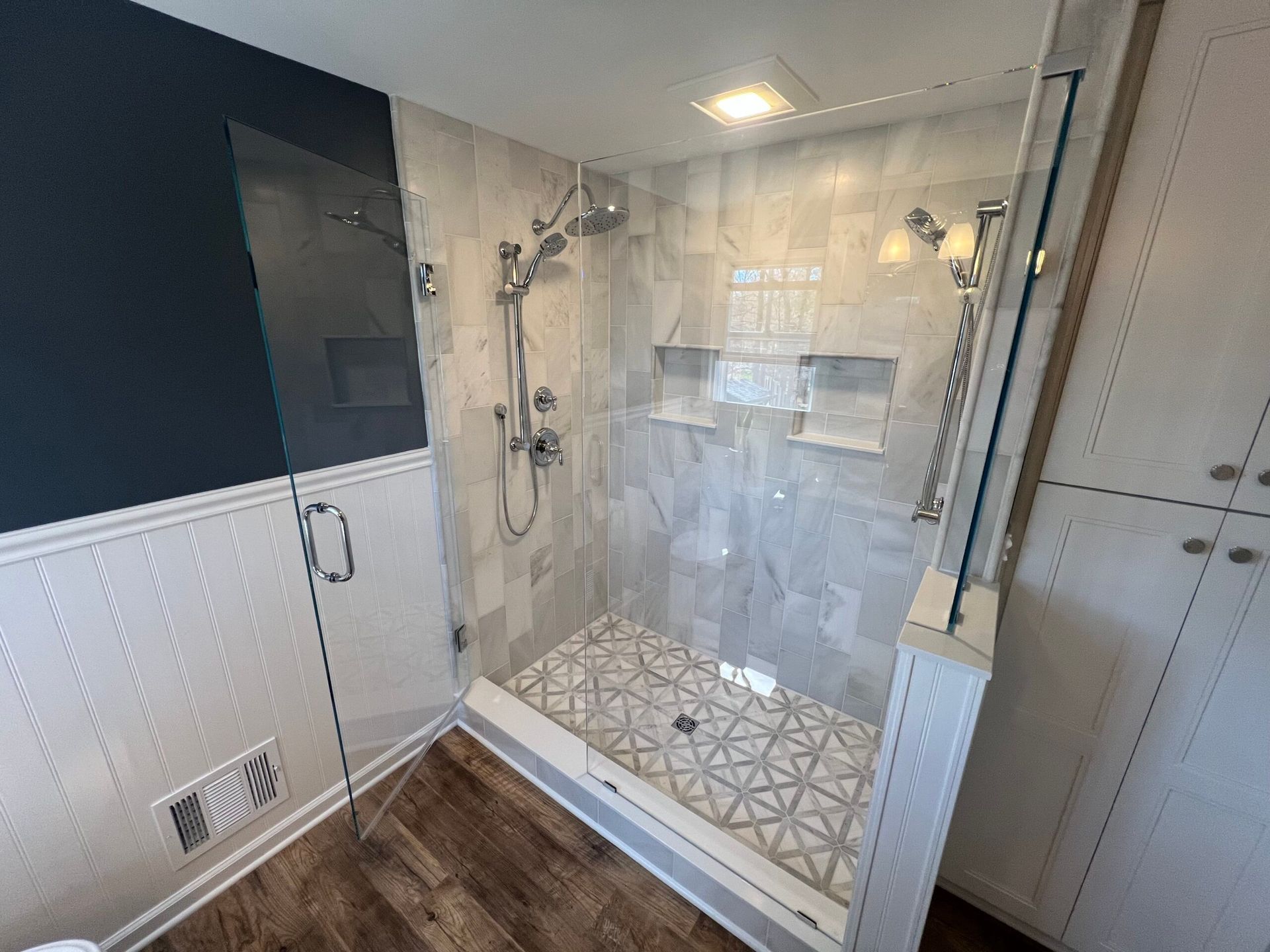 A tiled walk-in shower with a glass door, wall-mounted showerheads, recessed shelving, and a patterned tile floor.