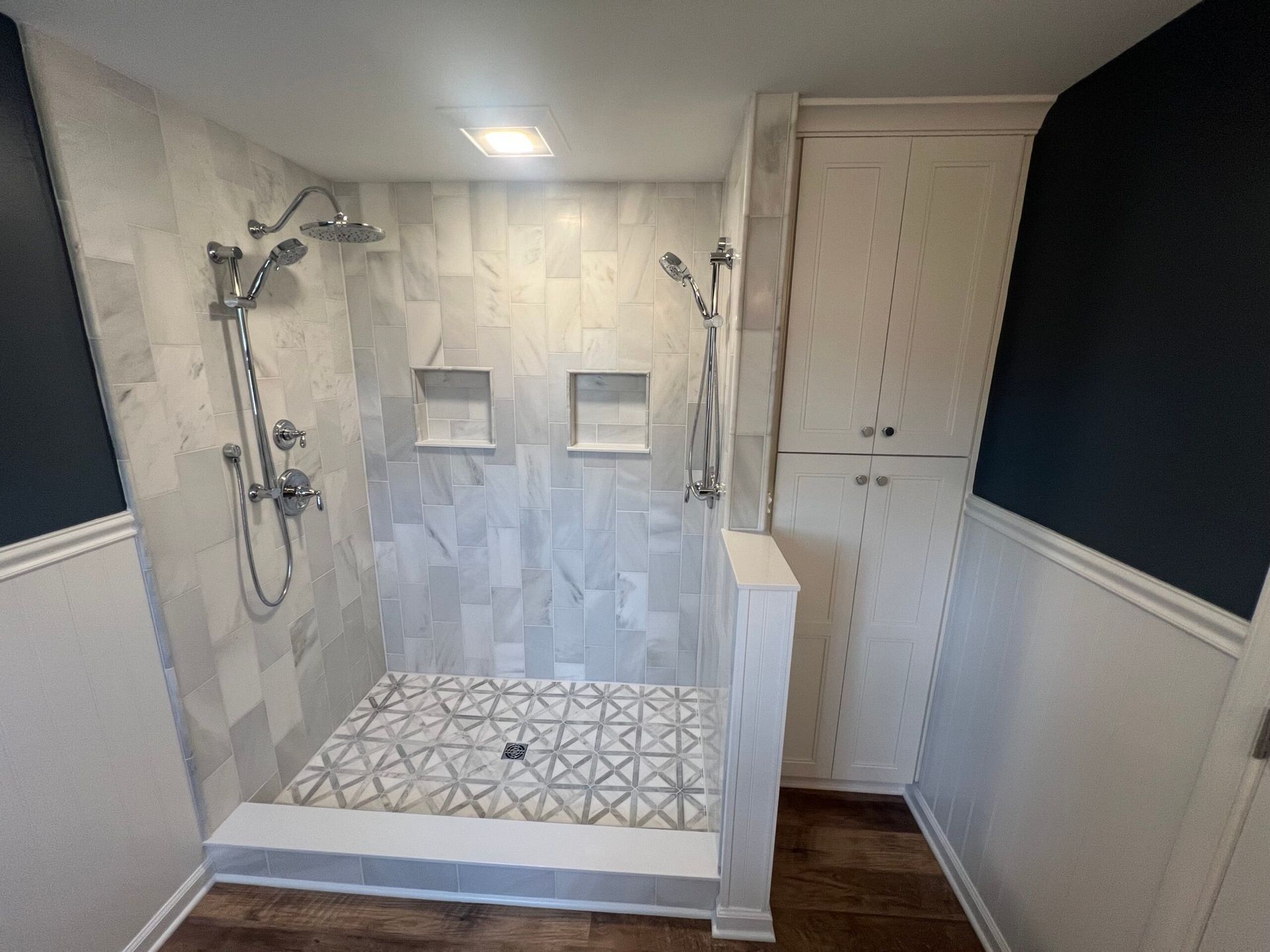 A walk-in shower with light grey patterned tiles, dual shower heads, built-in wall niches, and a tall white storage cabinet.