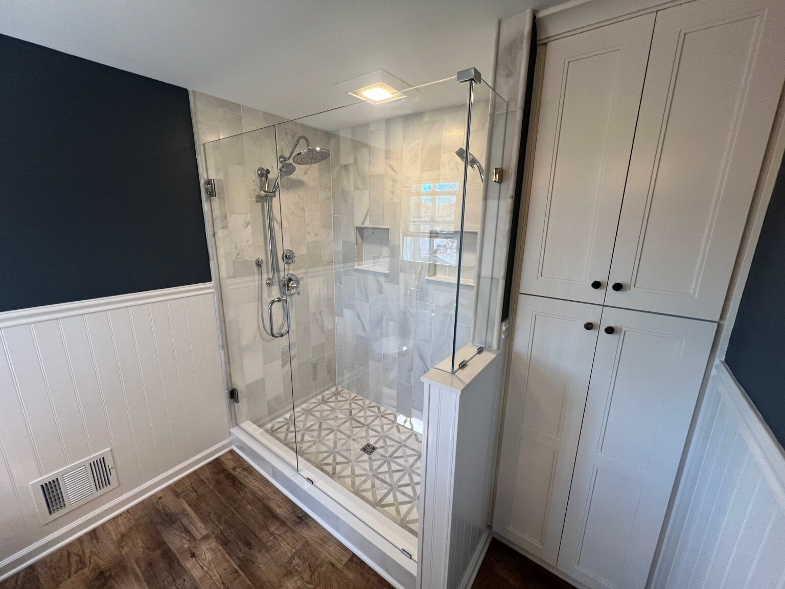 A modern walk-in shower with patterned floor tile, glass walls, and a white cabinet in a bathroom with dark blue walls.