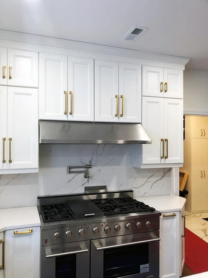 White kitchen cabinets with brass handles, a stainless steel stove with a range hood, and a marble backsplash.