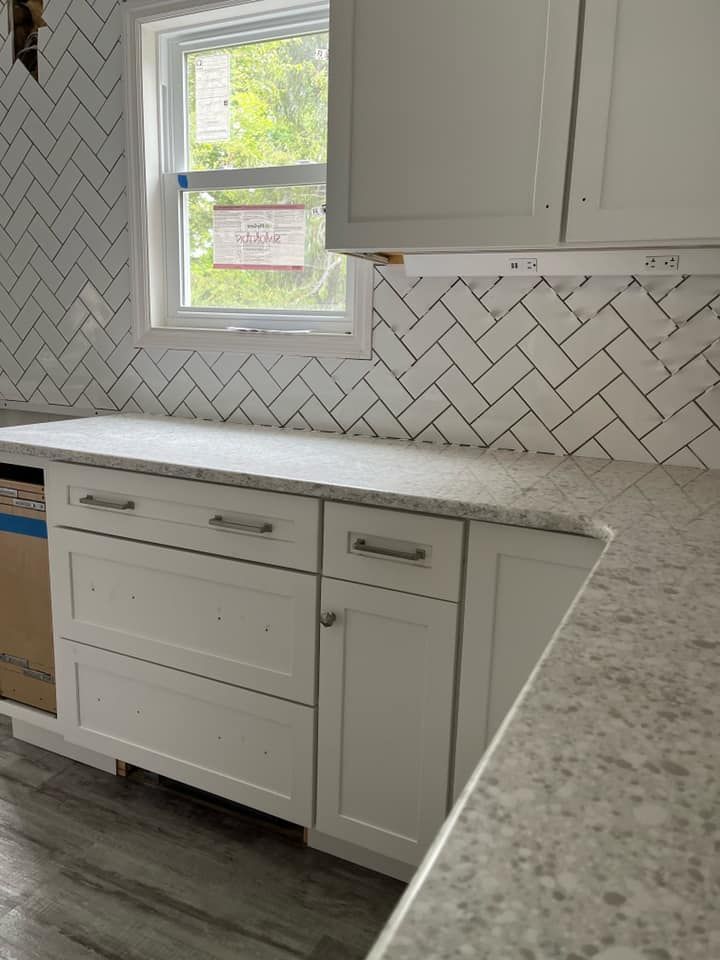 Bright kitchen with white cabinets, herringbone tile backsplash, and a window above the countertop.