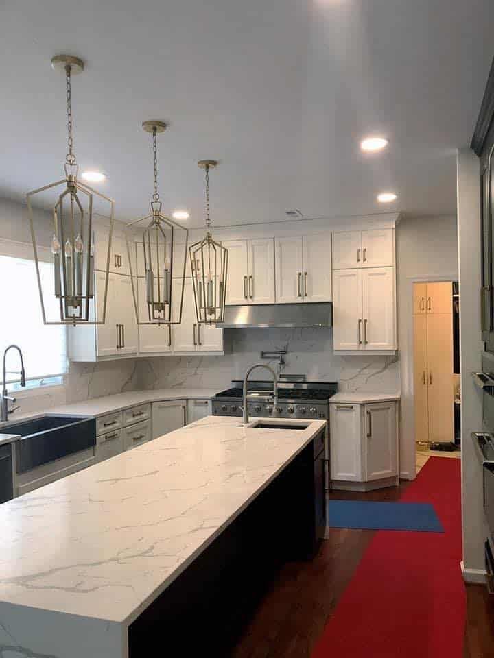 A modern kitchen with a white marble island, white cabinets, gold pendant lights, and a red runner rug on a wood floor.