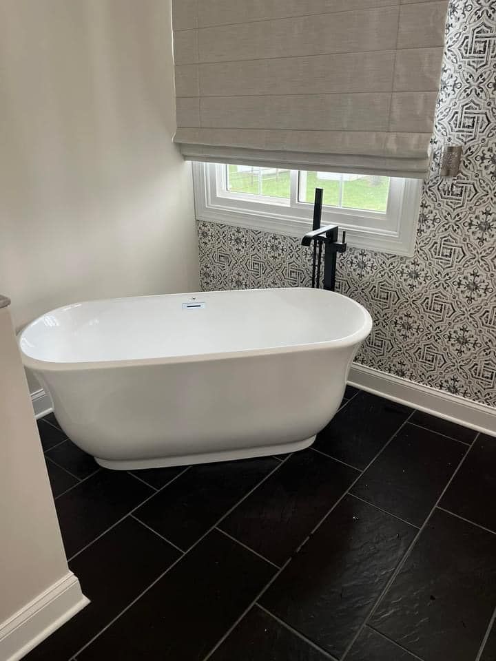 White freestanding bathtub in a bathroom with black tile flooring, patterned wall tiles, and a gray Roman shade.