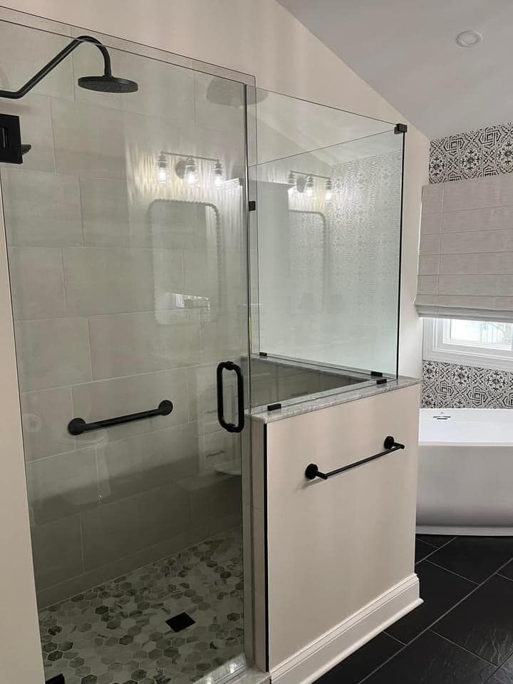 A walk-in shower with a glass enclosure, black hardware, a grab bar, and stone tile flooring, next to a white bathtub.