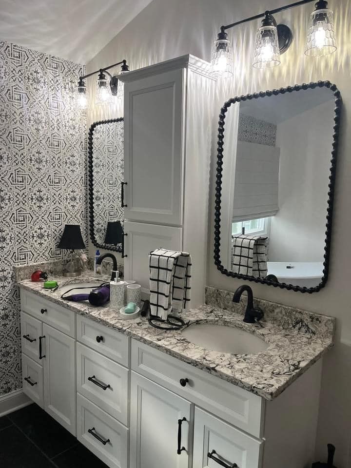 Double bathroom vanity with white cabinets, speckled stone countertops, framed mirrors, and patterned wallpaper.