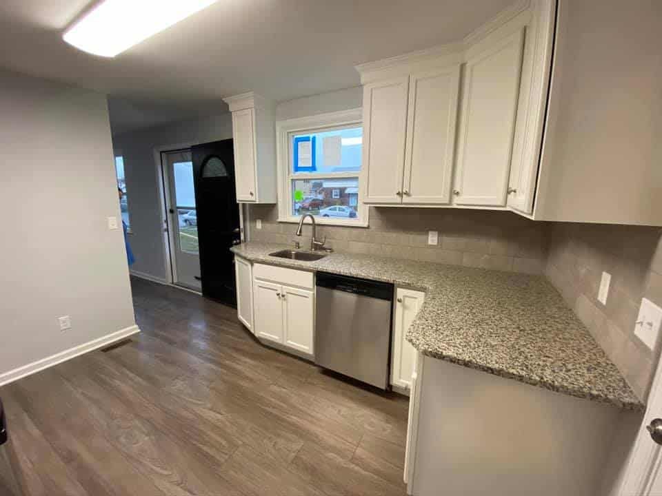 A kitchen with light wood flooring, white cabinets, a speckled granite countertop, stainless dishwasher, and a sink.