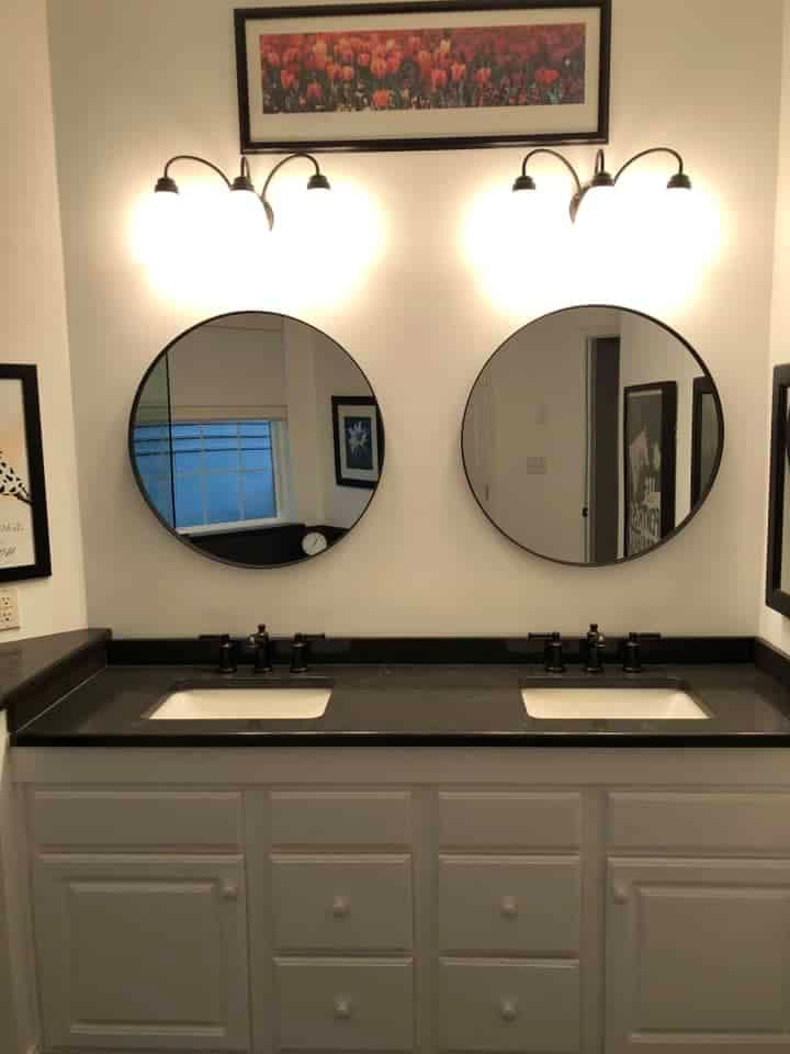A double bathroom vanity with white cabinets, a black countertop, two round mirrors, and two light fixtures overhead.