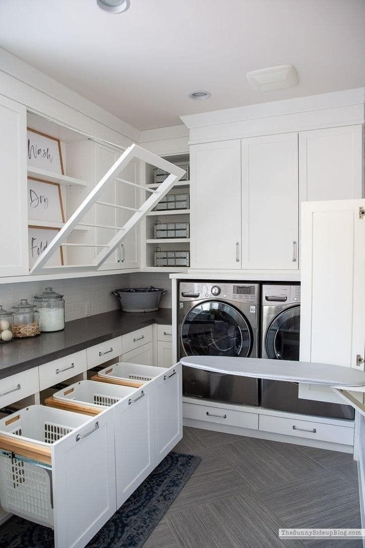 A bright laundry room with white cabinetry, a built-in ironing board, pull-out laundry hampers, and a fold-down drying rack.