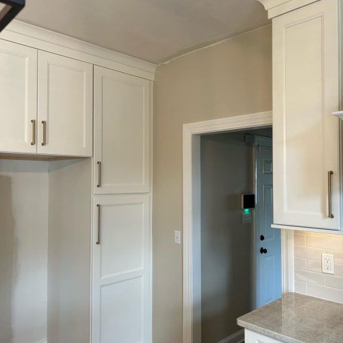 White kitchen cabinets and a tall pantry cabinet installed against a beige wall, next to a doorway to another room.