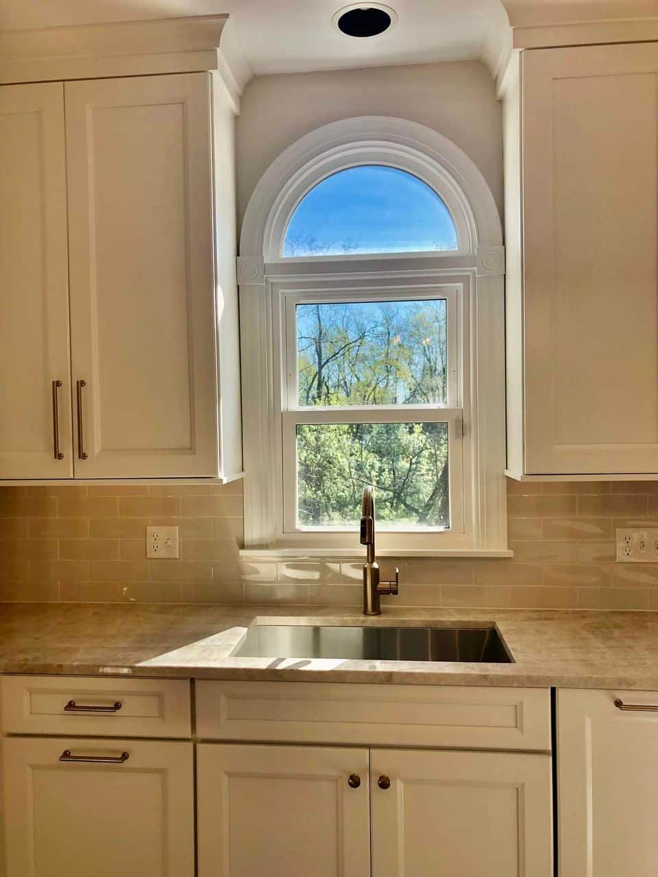 A kitchen sink with a chrome faucet positioned beneath an arched window, flanked by white cabinets and a light backsplash.