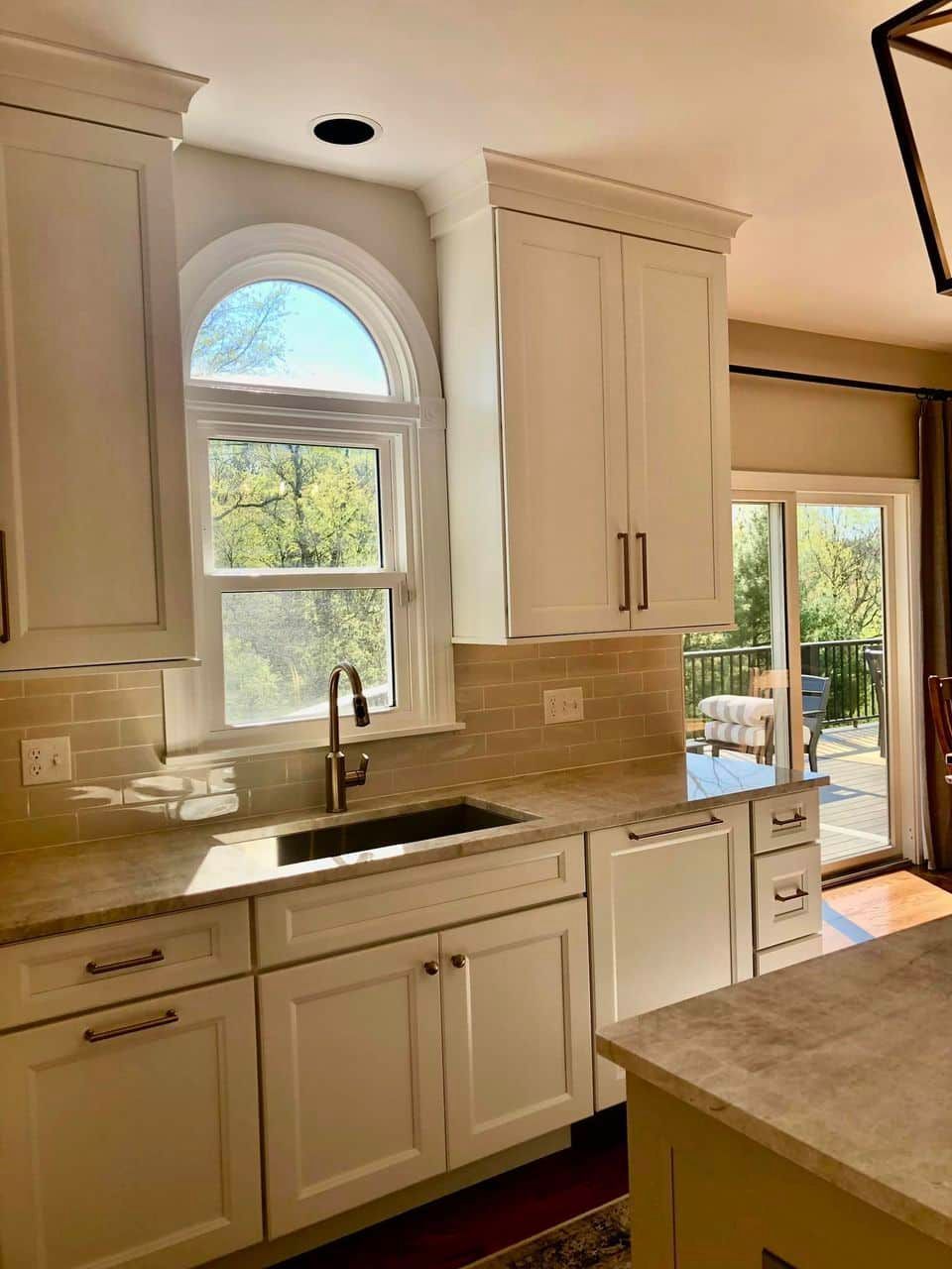 A white-cabinet kitchen with a sink, an arched window, and a sliding glass door leading to a deck.