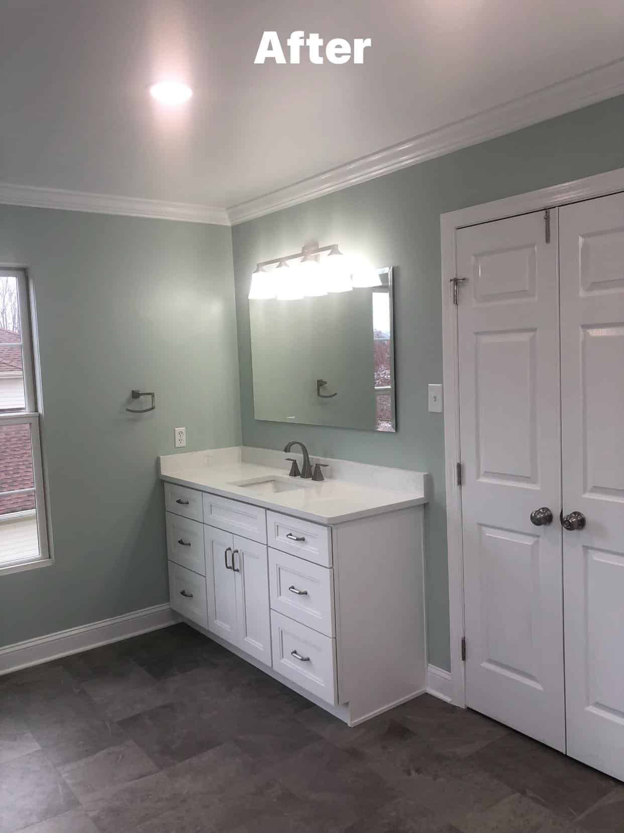 A bathroom with sage green walls, a white vanity with a mirror, and white double doors, featuring dark tile flooring.