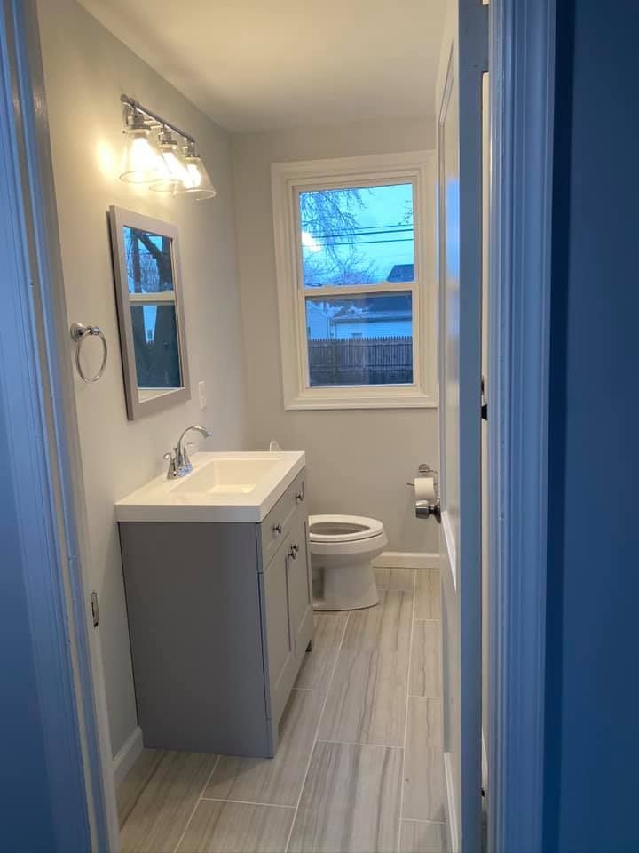 A bright bathroom featuring a grey vanity, a rectangular mirror with a light fixture above, and a toilet by a window.