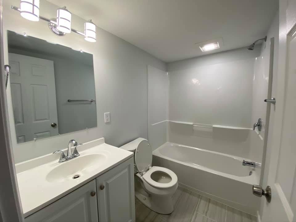 A well-lit, neutral-toned bathroom featuring a white vanity with a sink, a toilet, and a bathtub shower combo.