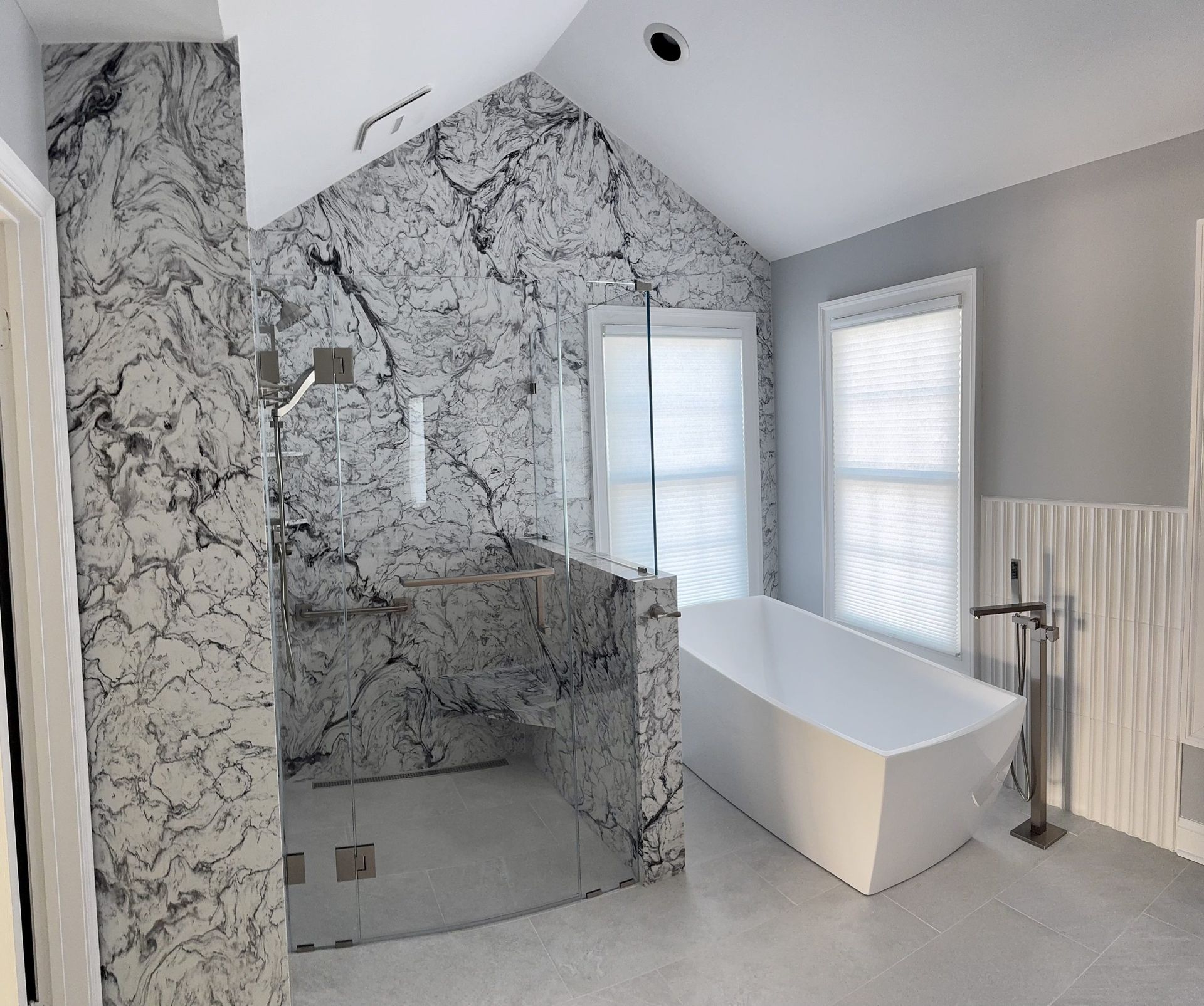 A luxury bathroom features a large, marble-tiled walk-in shower, a white freestanding tub, and soft gray walls.