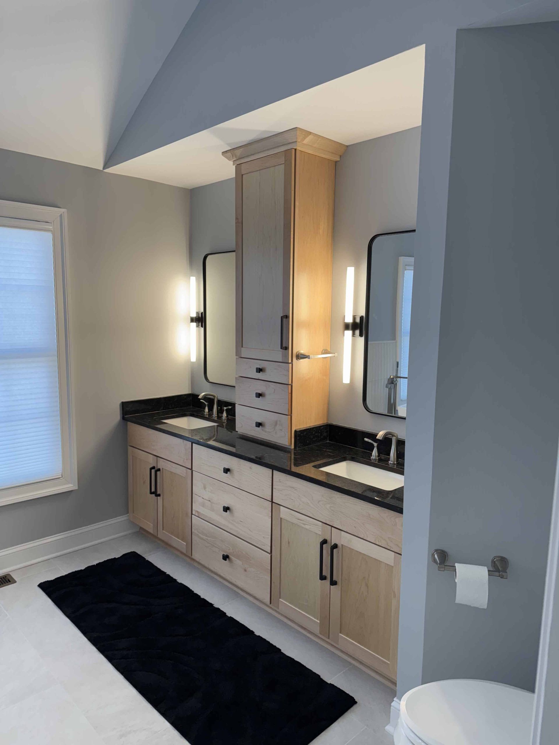 A bathroom vanity with wood cabinets, black countertops, twin mirrors, and a central tower, on a light tile floor.