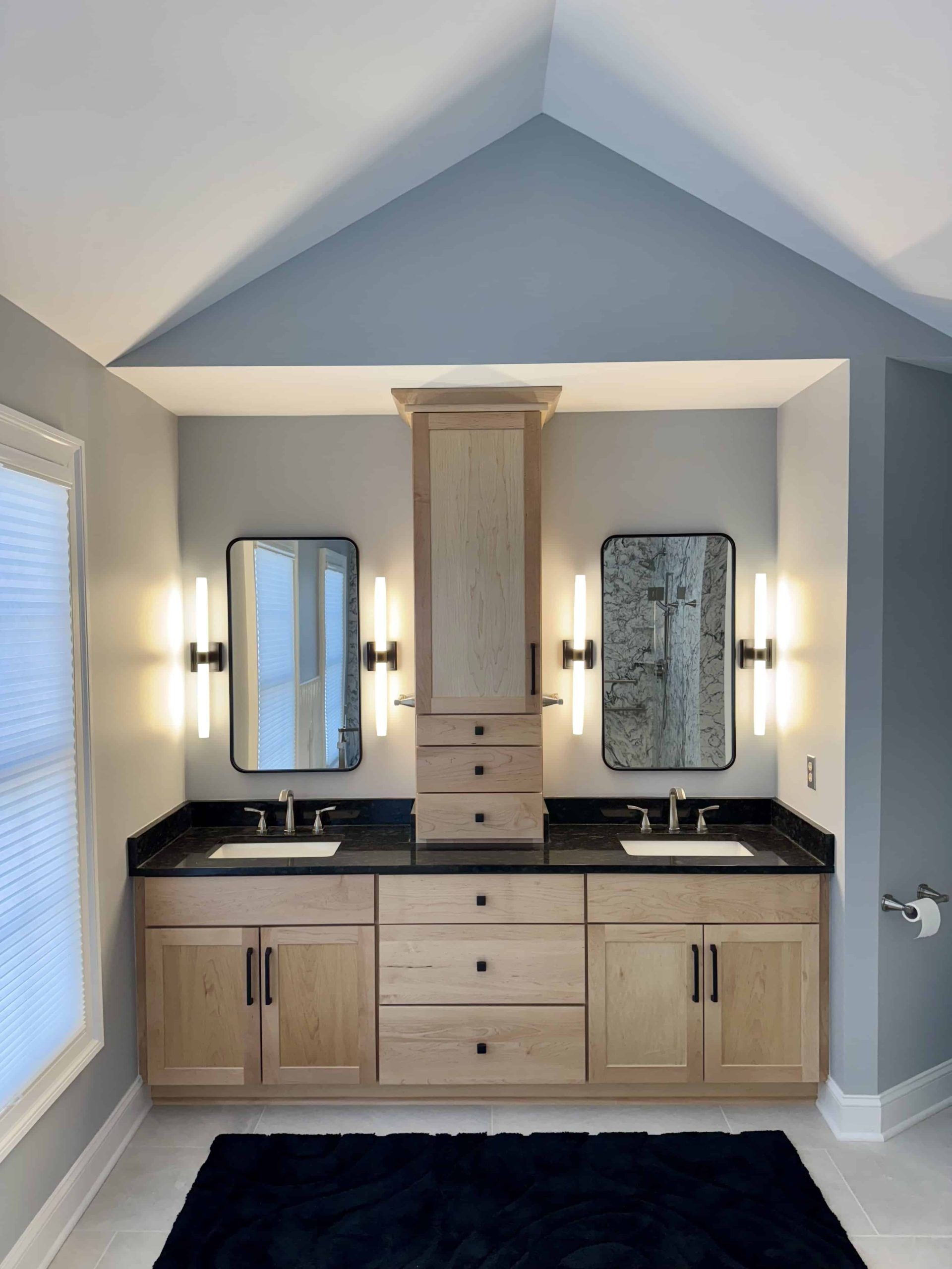 A bathroom double vanity with light wood cabinets, black countertops, twin mirrors, and wall sconces under a vaulted wall.