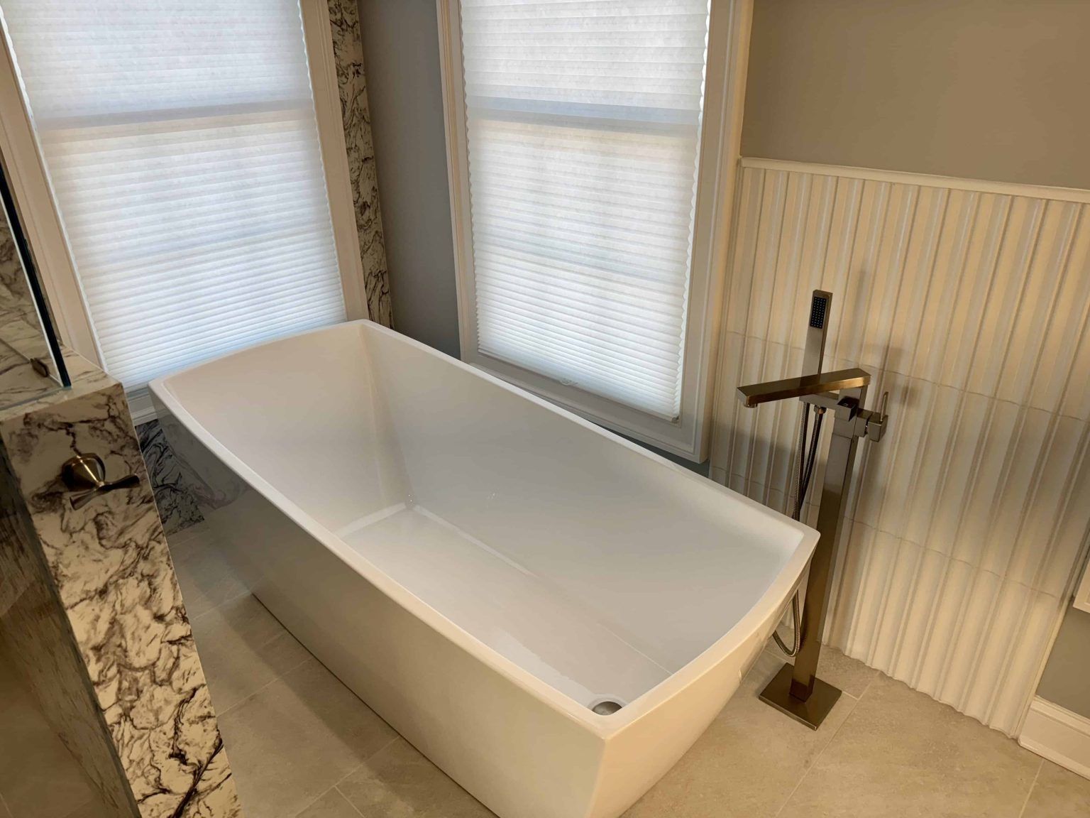 A white freestanding bathtub sits in a bathroom next to a tall brass floor-mounted faucet and windows with white blinds.