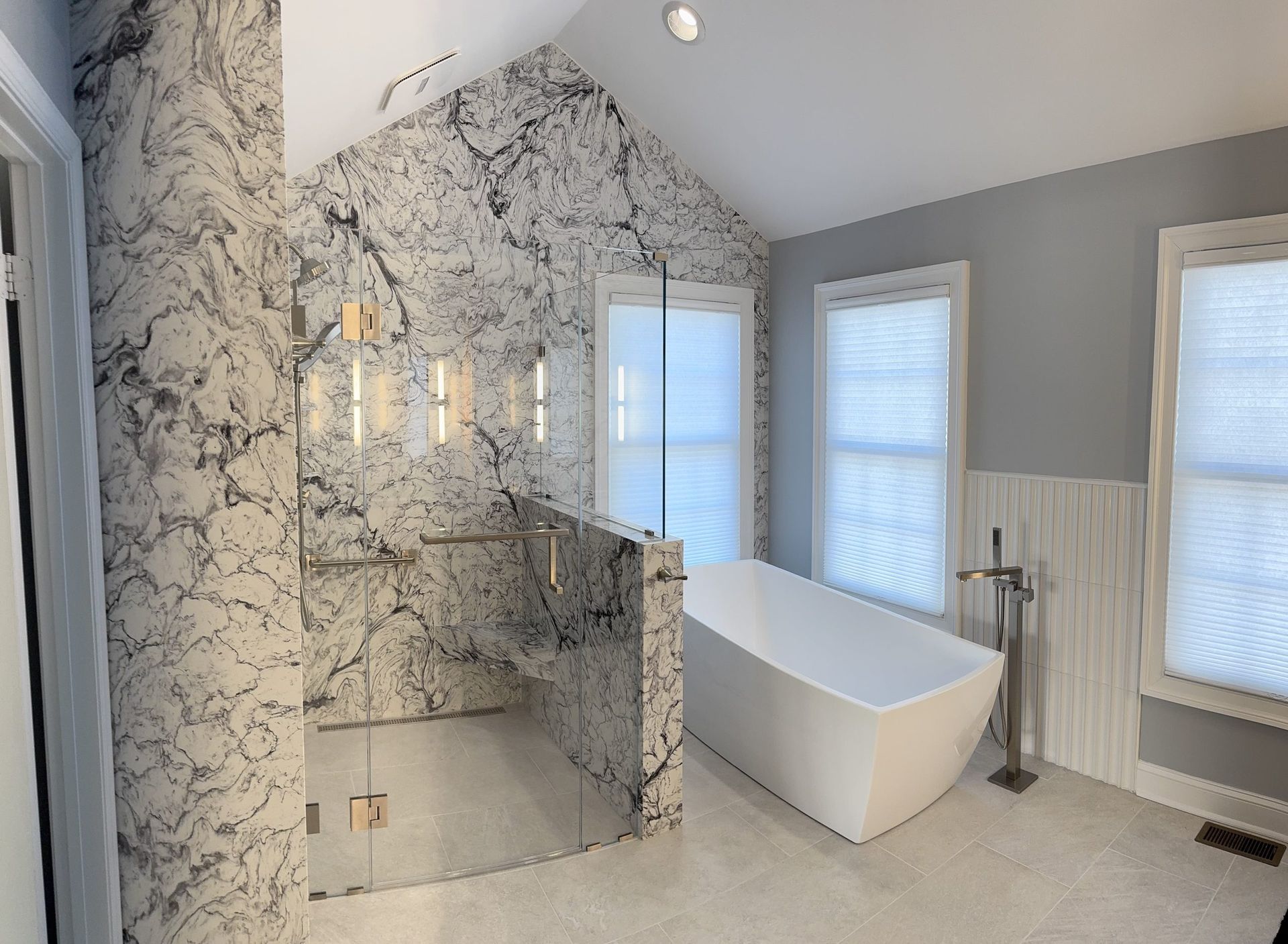 A modern bathroom featuring a white soaking tub, a marble-tiled walk-in shower with glass doors, and light gray walls.