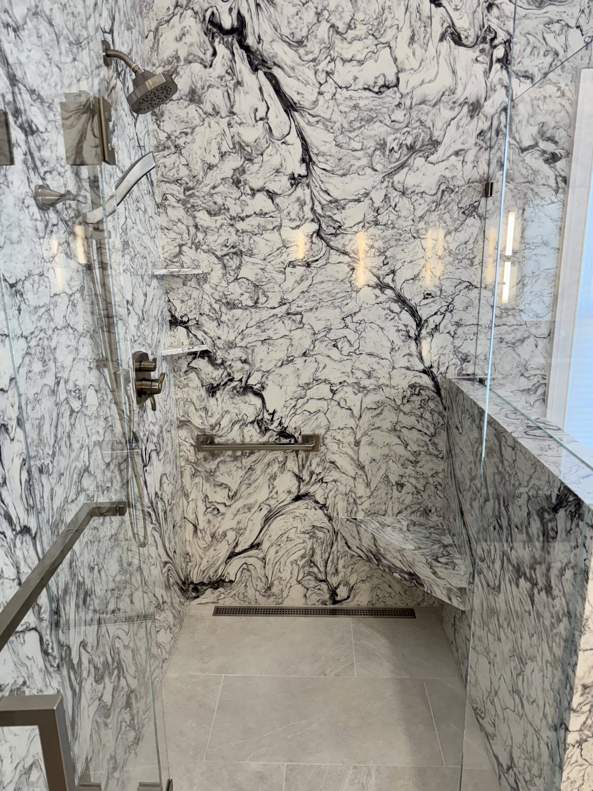 A modern walk-in shower with white and black marbled tile, a built-in stone bench, grab bars, and a linear floor drain.