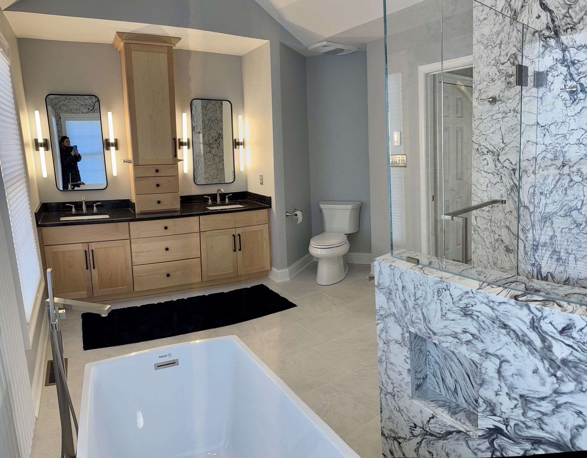 A luxury bathroom with light wood vanity, dual mirrors, a white standalone tub, and a marble-tiled shower enclosure.