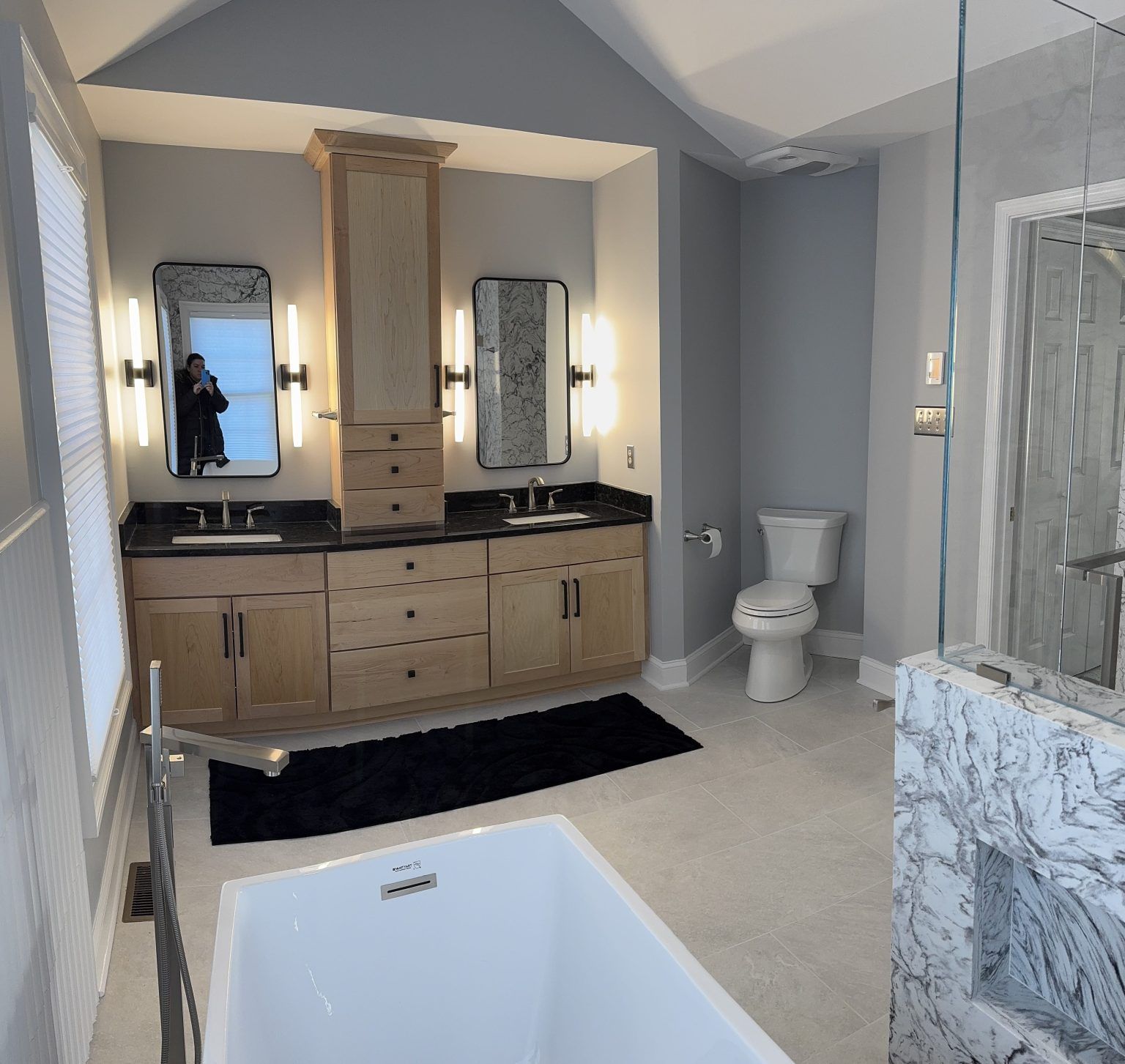 A modern bathroom with light wood dual vanities, white marble features, a freestanding tub, and a toilet.