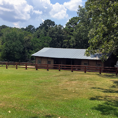 Wooden barn with gray roof behind a red fence in a grassy field, surrounded by trees and blue sky.