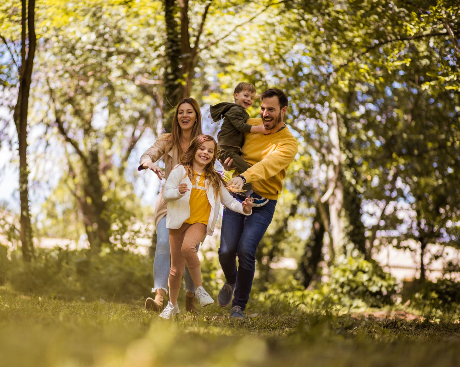 Family of four in a sunny park, smiling. Mom on scooter, dad holding child, girl running. Green grass and trees.