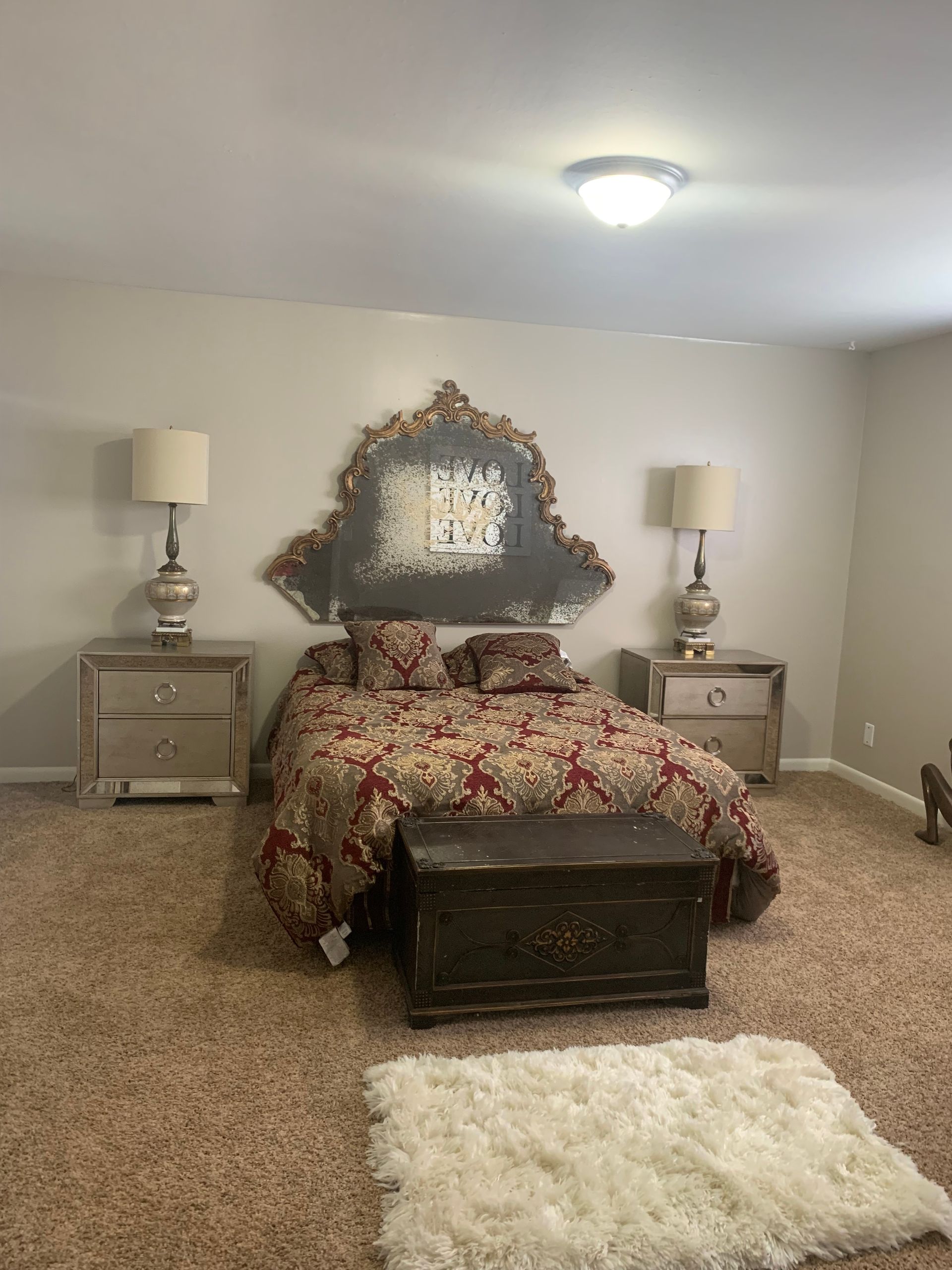 Bedroom with decorative headboard, bed, nightstands, lamps, and a chest. Beige walls, carpet, and a fluffy rug.