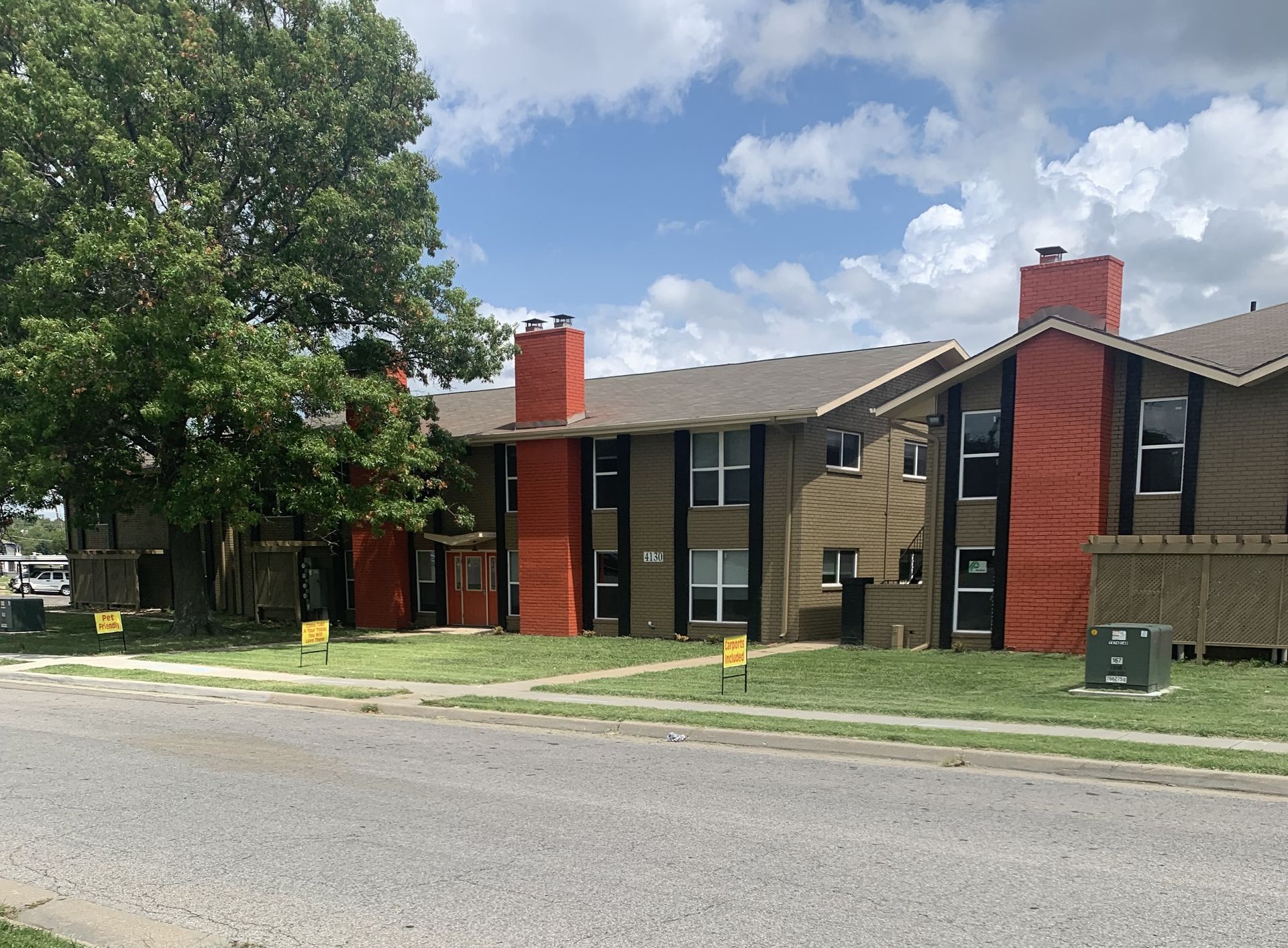 Two-story brown apartment buildings with orange chimneys and accents, on a green lawn beside a street.