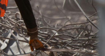 Worker's gloved hand using a tool amid twisted metal and debris, possibly after a disaster.